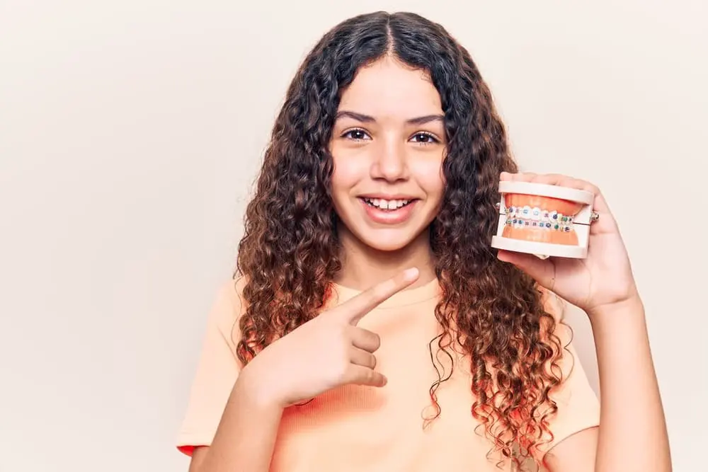 Girl with curly hair smiling and pointing at a dental model with braces, wearing a peach shirt, highlighting the journey to a perfect smile. Braces cost considerations included in Vacaville, CA.