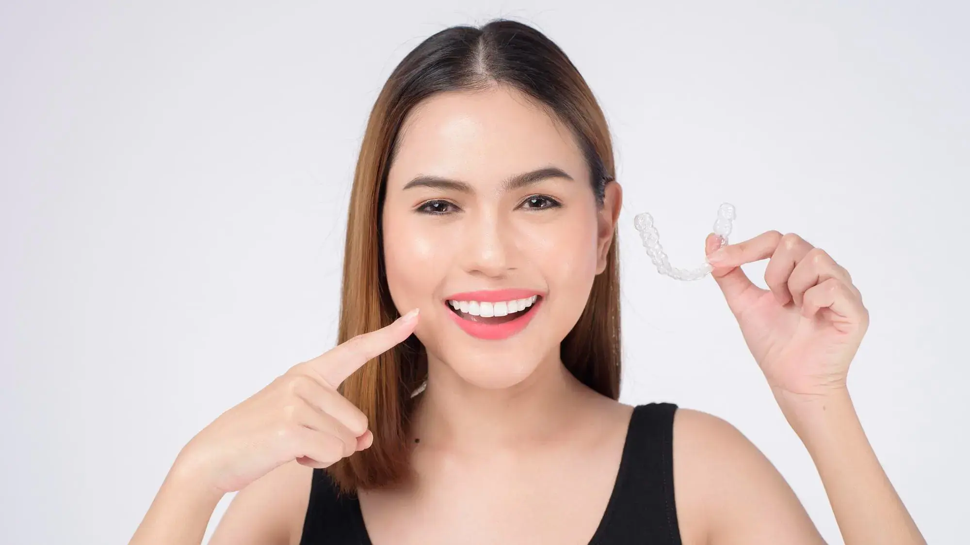 Woman smiling, holding a clear dental aligner in one hand and pointing to her teeth with the other at Natalie Yang Orthodontics in Vacaville, CA. Woman smiling, holding a clear dental aligner in one hand and pointing to her teeth with the other at Natalie Yang Orthodontics in Vacaville, CA.
