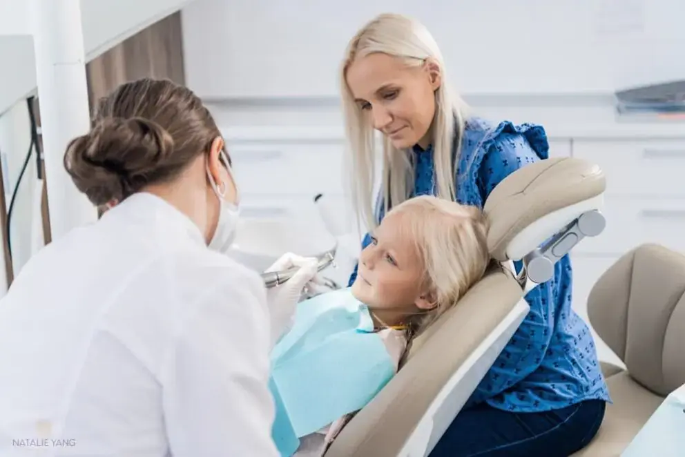 A dentist examines a young child in a dental chair while an adult woman sits nearby, observing in a bright dental office. A dentist examines a young child in a dental chair while an adult woman sits nearby, observing in a bright dental office.