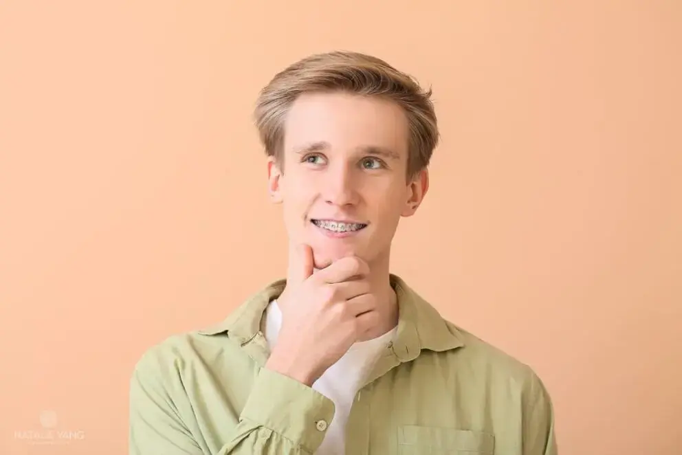 Young man with braces wearing a light green shirt poses against a peach-colored background, resting his chin on his hand and looking slightly upward. Young man with braces wearing a light green shirt poses against a peach-colored background, resting his chin on his hand and looking slightly upward.