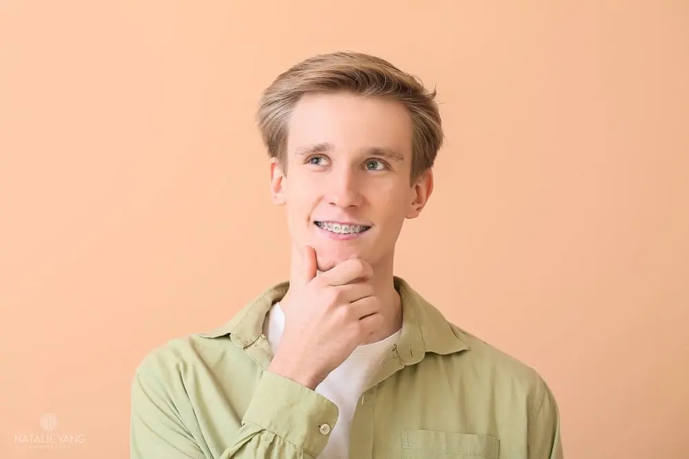 Young man with braces wearing a light green shirt poses against a peach-colored background, resting his chin on his hand and looking slightly upward after understanding how long do braces take in Vacaville, CA..