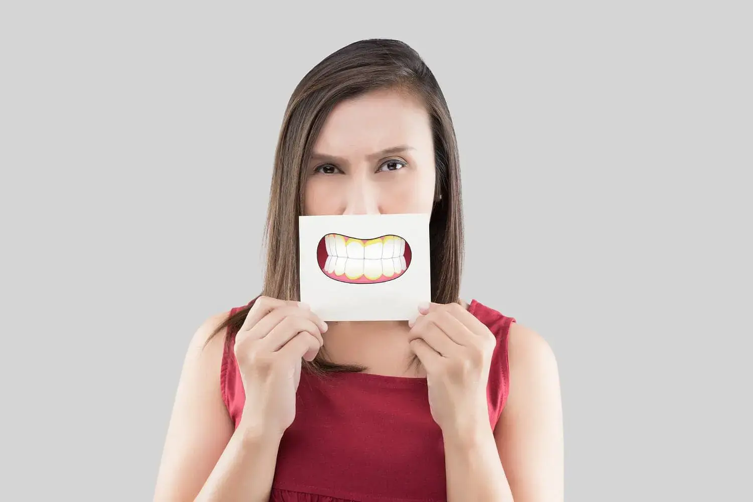 A woman in a red sleeveless top stands against a plain gray background, holding a paper with a cartoon underbite smile over her face—a playful image from Natalie Yang Orthodontics Orthodontics in Vacaville, CA. A woman in a red sleeveless top stands against a plain gray background, holding a paper with a cartoon underbite smile over her face—a playful image from Natalie Yang Orthodontics Orthodontics in Vacaville, CA.