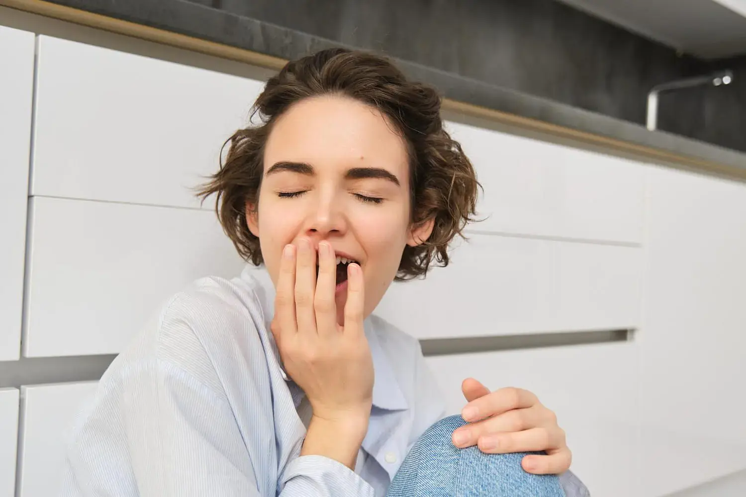 At Natalie Yang Orthodontics Orthodontics in Vacaville, CA, a person with short brown hair is seen sitting before white cabinets, yawning with eyes closed and hand covering their mouth—demonstrating the typical posture of a mouth breather. At Natalie Yang Orthodontics Orthodontics in Vacaville, CA, a person with short brown hair is seen sitting before white cabinets, yawning with eyes closed and hand covering their mouth—demonstrating the typical posture of a mouth breather.