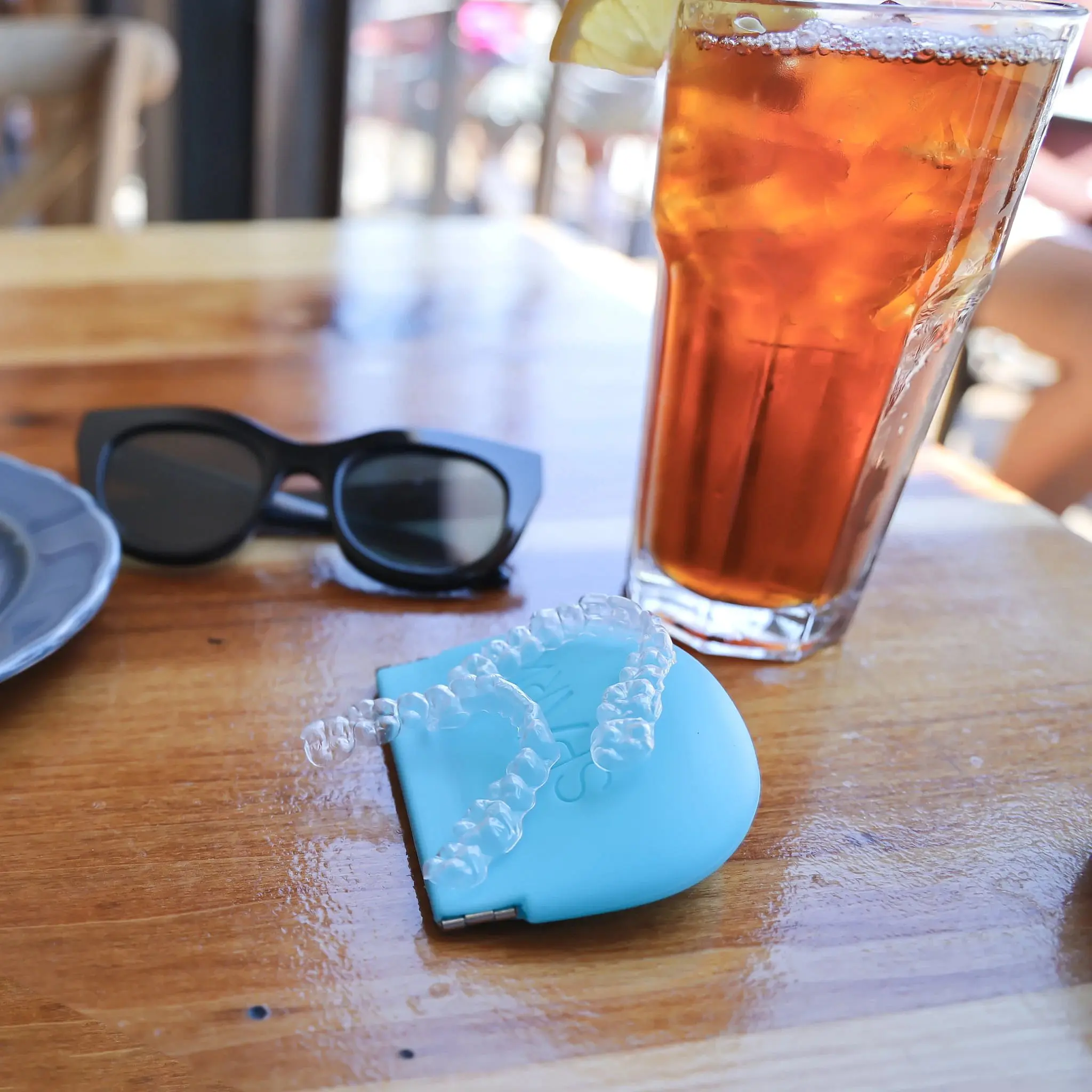 On a wooden table in Vacaville, CA, a pair of clear aligners in a blue case, black sunglasses, and a glass of iced tea with lemon are arranged, highlighting Natalie Yang Orthodontics.