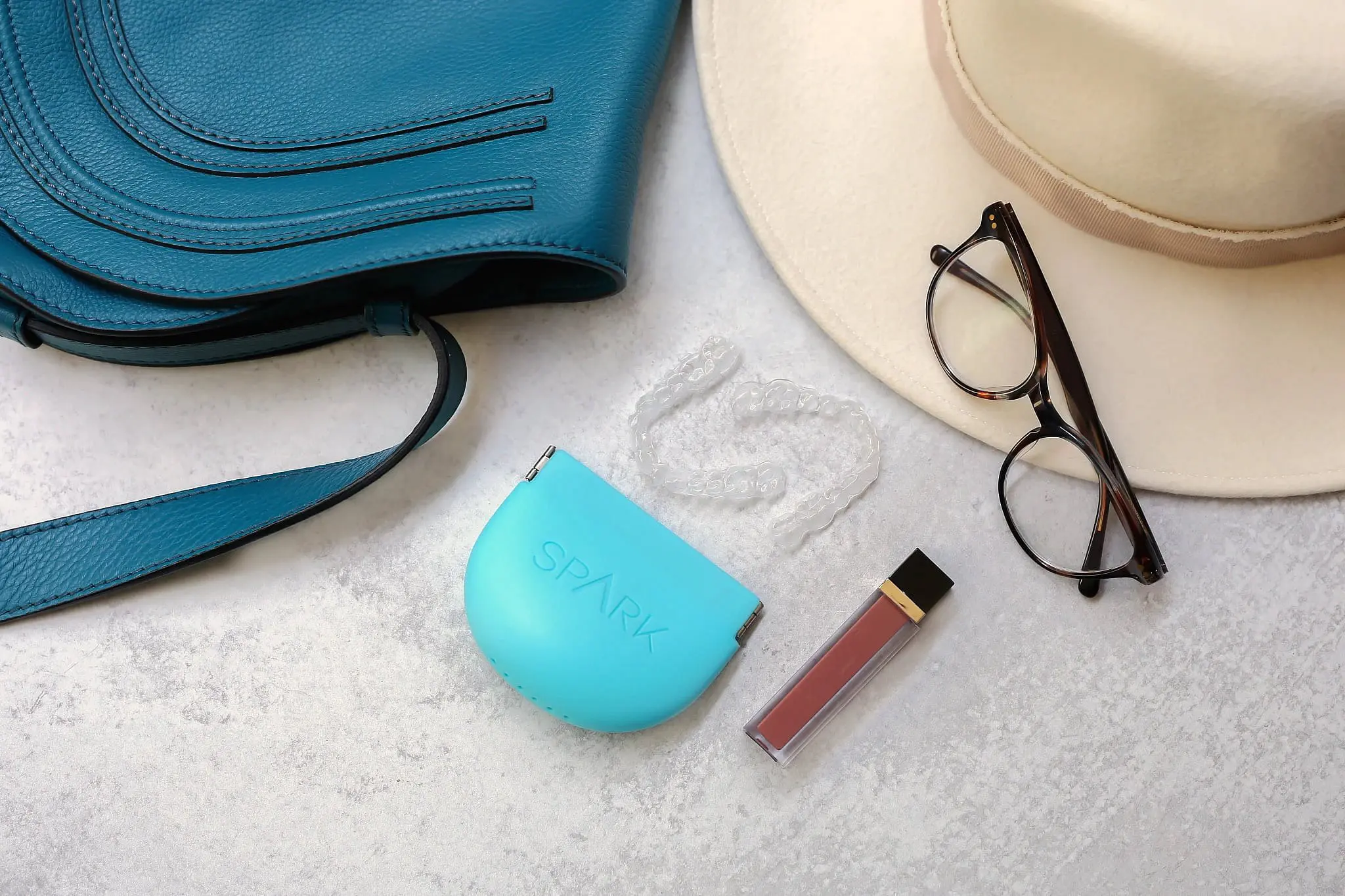 Featured in this collection for Natalie Yang Orthodontics in Vacaville, CA, a blue handbag, beige hat, black-rimmed glasses, a tube of lip gloss, and a clear dental aligner with its blue Spark case are neatly arranged on a light surface.