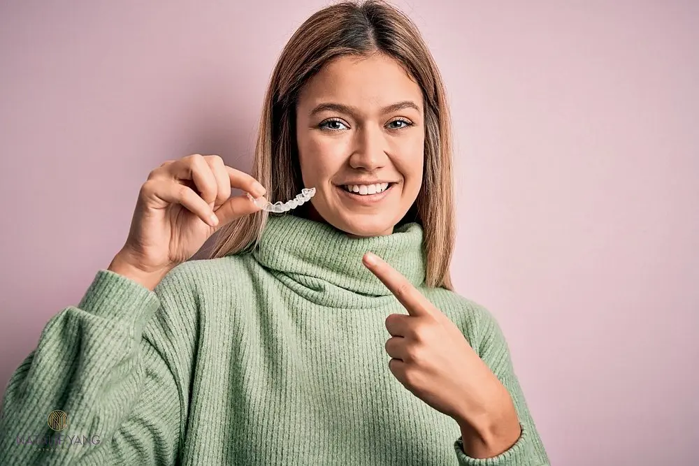 Young woman in a green sweater holding and pointing at Invisalign clear aligners after understanding Does Insurance Cover Invisalign in Vacaville, CA