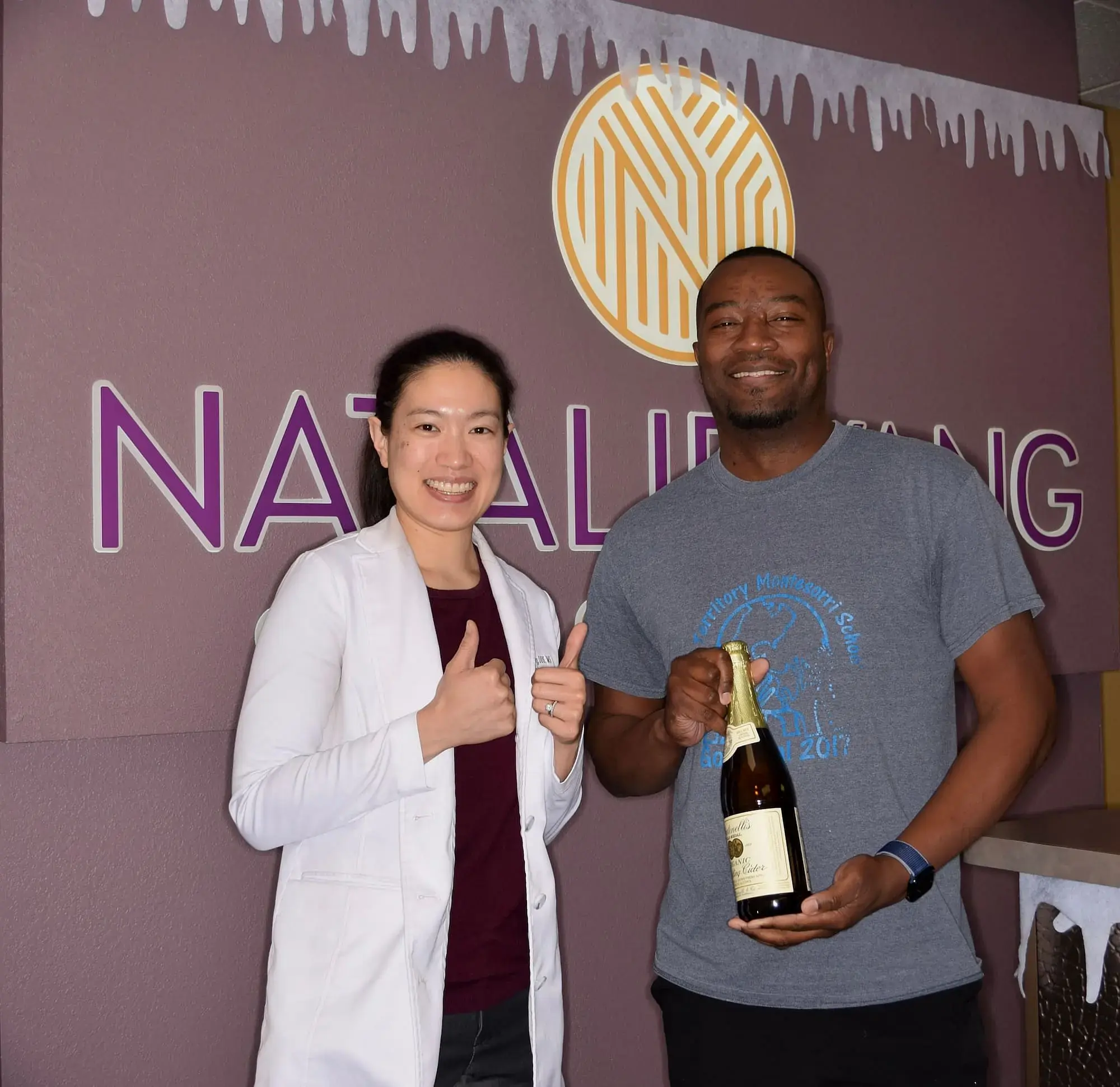Smiling pair giving thumbs up in front of wall sign at Natalie Yang Orthodontics in Vacaville, CA, celebrating overbite treatment success. Smiling pair giving thumbs up in front of wall sign at Natalie Yang Orthodontics in Vacaville, CA, celebrating overbite treatment success.