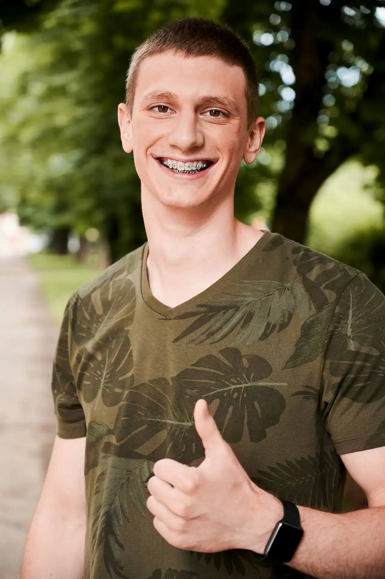 Smiling young man with adult braces in a green leaf shirt gives a thumbs up outdoors at Natalie Yang Orthodontics in Vacaville, CA. Smiling young man with adult braces in a green leaf shirt gives a thumbs up outdoors at Natalie Yang Orthodontics in Vacaville, CA.
