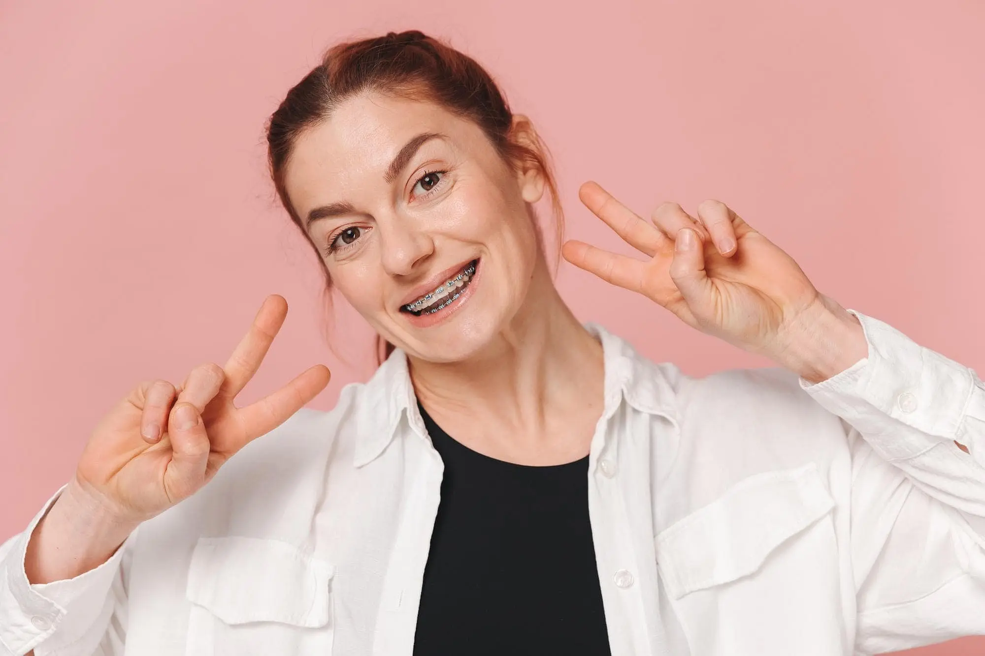 Smiling woman with metal braces shows peace signs against a pink backdrop at Natalie Yang Orthodontics in Vacaville, CA.