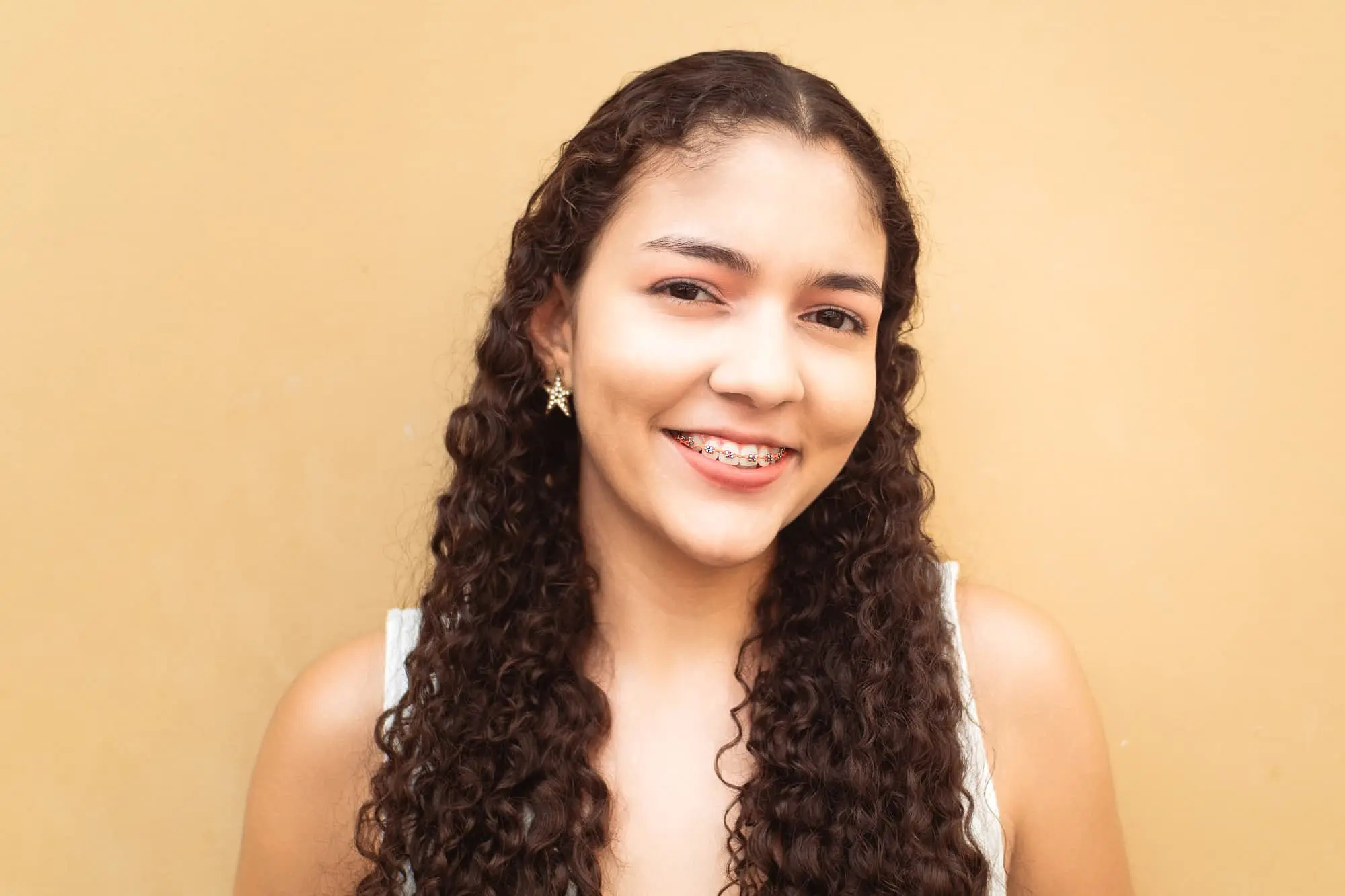 Smiling young woman with long curly hair and metal braces at Natalie Yang Orthodontics in Vacaville, CA, beige background.