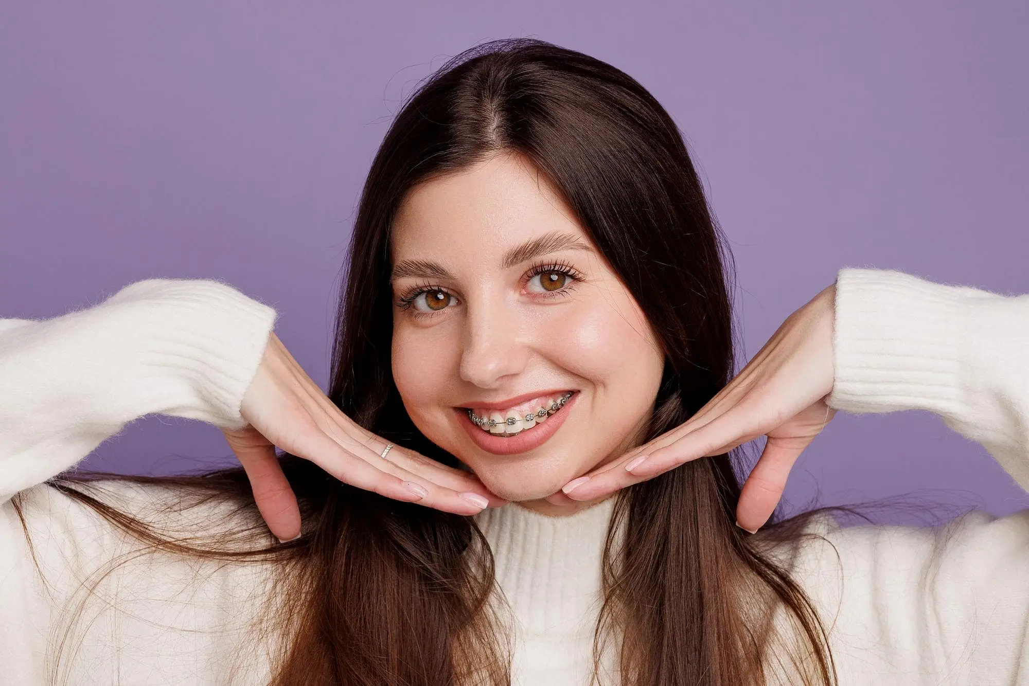 A young woman with metal braces smiles, hands framing her face, for Natalie Yang Orthodontics in Vacaville, CA.